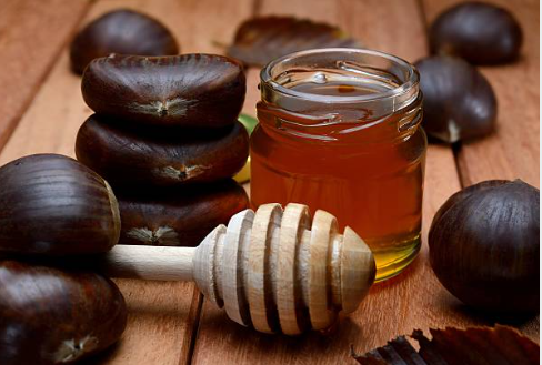 pot de miel de châtaignier posé sur une table, avec une cuillère de miel en bois à côté, et des châtaignes marrons disposées autour.