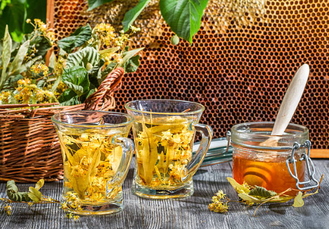 Photo d'un pot de miel de tilleul avec une cuillère en bois plongée à l'intérieur, prête à être utilisée pour sucrer le thé. À côté du pot, plusieurs tasses de thé sont disposées.