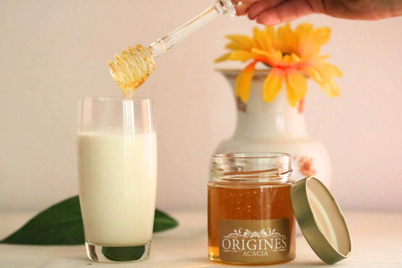 notre pot de miel d'acacia posé sur une table, avec une personne qui utilise une cuillère pour verser doucement du miel dans un verre de lait, créant une harmonie de saveurs délicieuse et réconfortante.